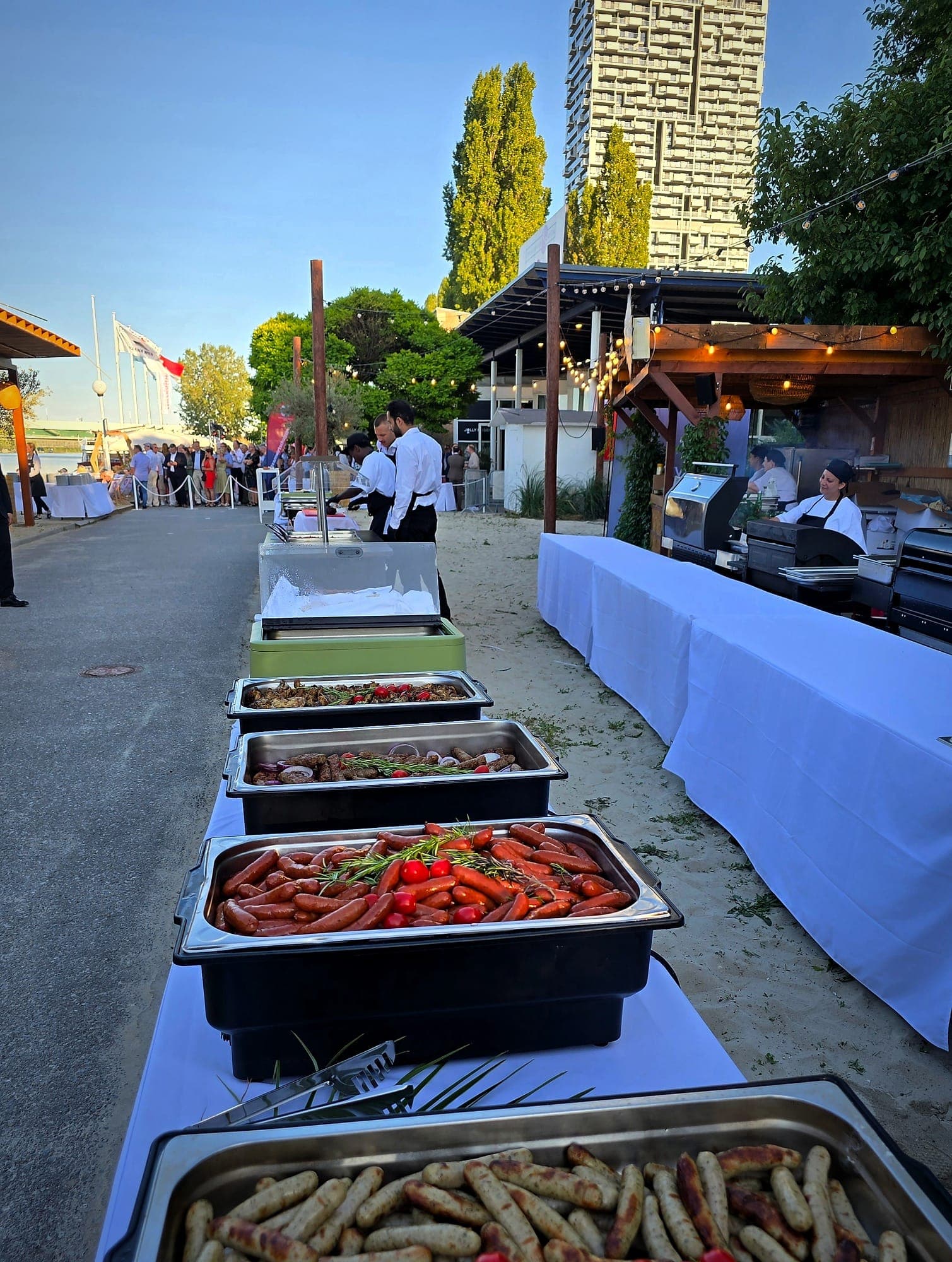Grill-Buffet von charlyfresh im Freien — Bratwürste und Grillgut in Chafing Dishes, Grillstation im Hintergrund